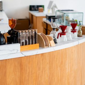 A modern coffee shop counter featuring a sleek wooden design. There are glass containers with coffee beans, brewing equipment like a pour-over setup, and a coffee grinder